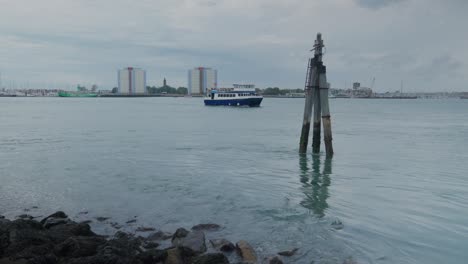 Wide-shot-of-seawater-hitting-rocks-and-post-on-a-calm-day-in-the-harbour,-a-boat-is-passing-by-with-council-estates-in-the-background