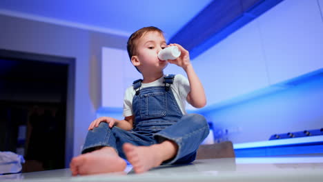 adorable caucasian toddler in jeans romper and white t-shirt sits on the table and drinks from bottle. low angle view at the kid having a snack.