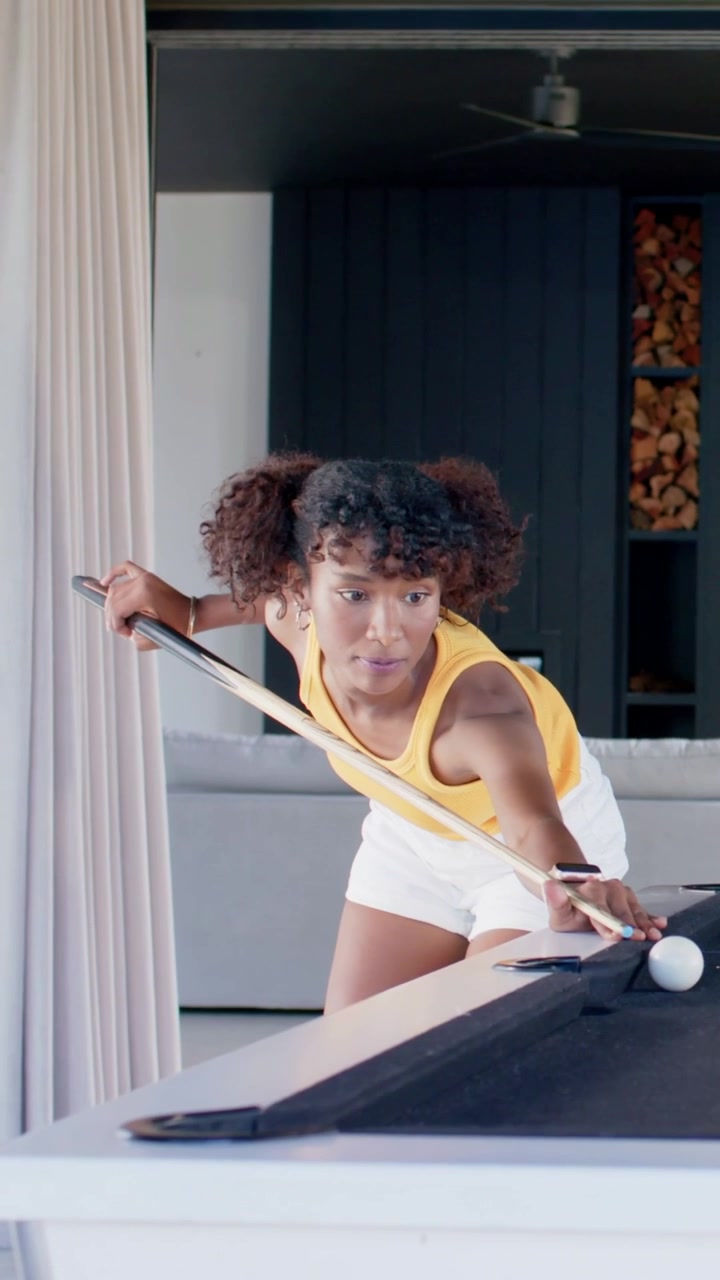 Vertical video: Approaching pool table woman yellow top shorts aiming cue hitting cue ball at home