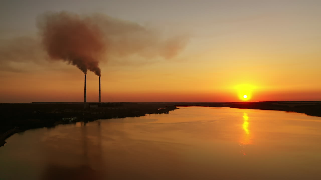 Industrial plant near the evening river. Dirty smoke from pipes goes in the air against the setting sun. Silhouette of a factory near the lake on orange sky background.