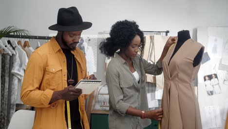 hombre y mujer jóvenes, diseñadores de ropa, sonriendo a la cámara mientras toman medidas del vestido con cinta métrica