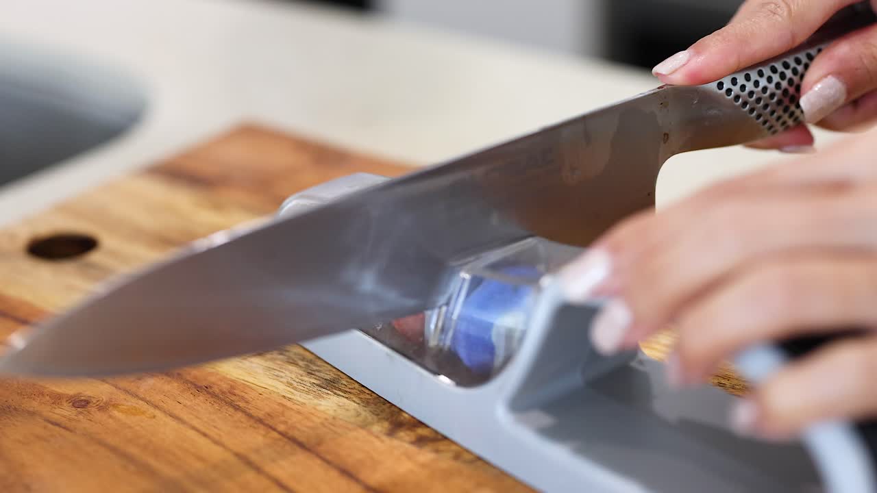 A person sharpens a knife using a manual sharpener on a wooden countertop with natural lighting
