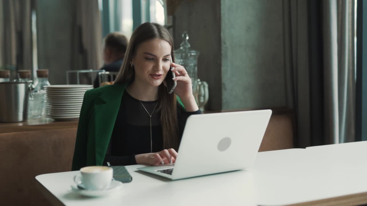 Smuk ung kvinde, en freelancer, sidder på en stilfuld restaurant klædt i forretningstøj, taler i telefon og arbejder på sin bærbare computer