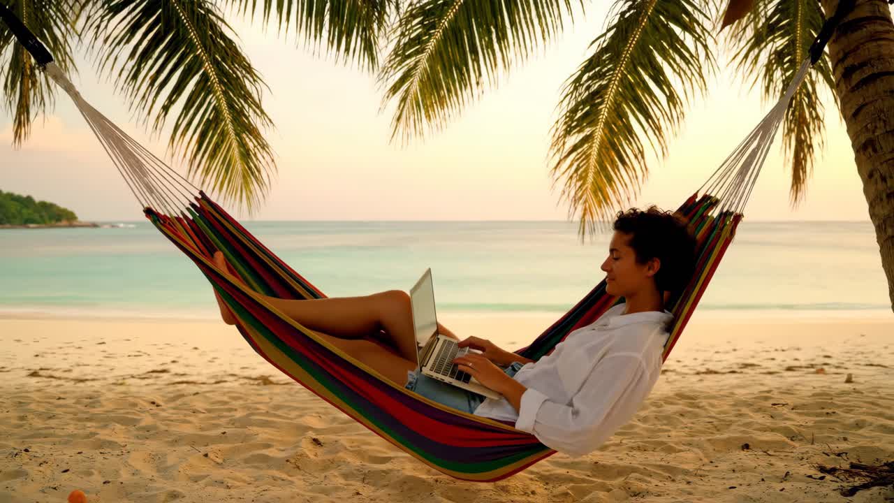 Woman relaxing in a hammock on a tropical beach while working on her laptop