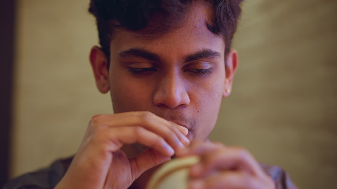Young man in close-up portrait holding drink near lips while gazing with focused, thoughtful expression in warm light setting, conveying emotion, introspection, and deep engagement in quiet indoor atmosphere