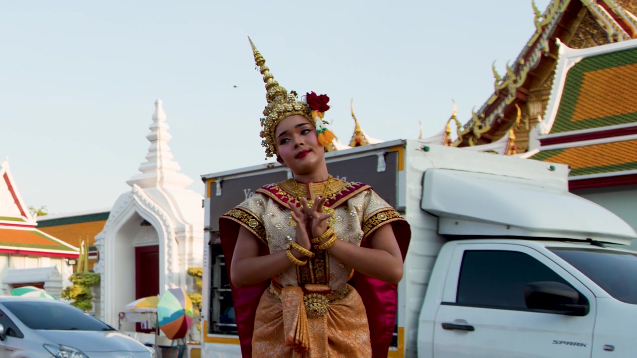 En danser i udsmykket guldkostume udfører klassiske thailandske bevægelser uden for et historisk tempel