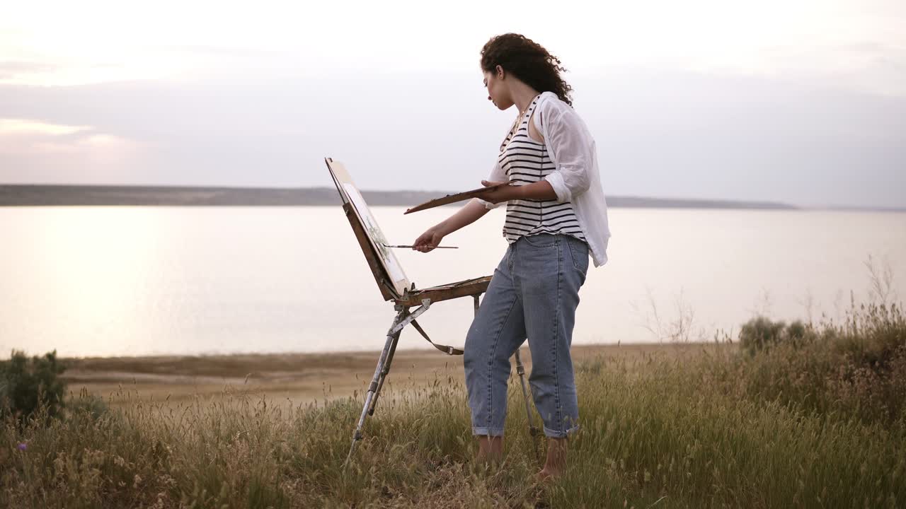 Full length footage of a beautiful girl in white shirt and jeans working outdoors. Paints using an easel and oil paints on the meadow in front the lake. Wind blowing, morning