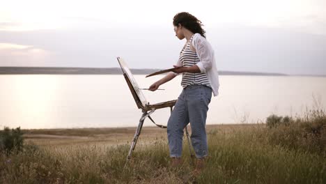 Imágenes-Completas-De-Una-Hermosa-Chica-Con-Camisa-Blanca-Y-Jeans-Trabajando-Al-Aire-Libre.-Pinta-Con-Caballete-Y-Pinturas-Al-óleo-En-El-Prado-Frente-Al-Lago.-Viento-Que-Sopla,-Mañana