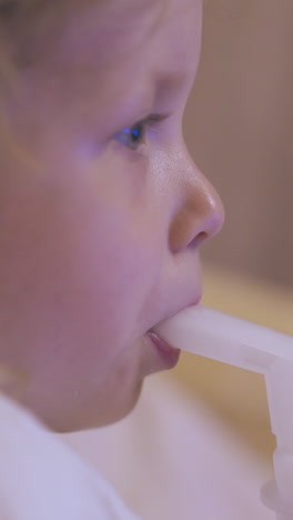 unhappy little blonde girl has procedure with plastic inhaler lying in bed on blurred background at home extreme close view