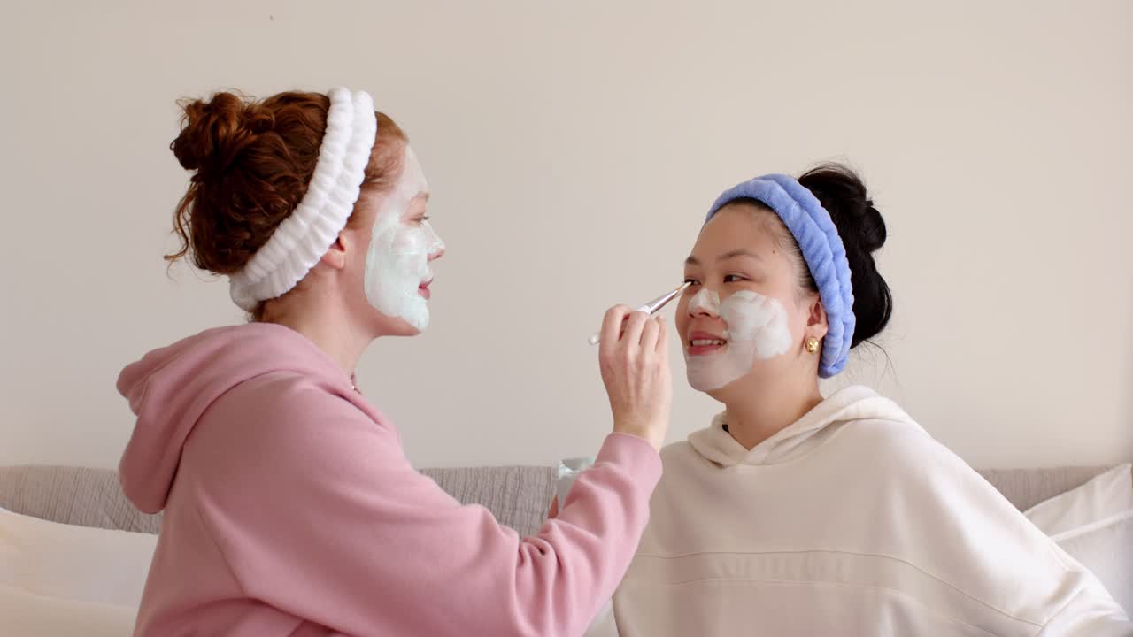 Dua teman wanita di sofa, wanita di sebelah kiri mencelupkan kuas, mengoleskan masker ke teman wanitanya yang orang Asia untuk mempererat ikatan