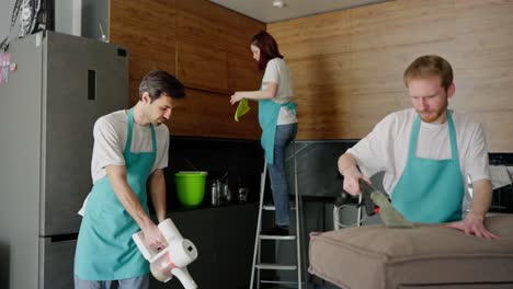 Side-view-of-a-confident-group-of-two-cleaning-guys-and-a-brunette-girl-in-a-white-T-shirt-and-blue-apron-cleaning-in-a-modern-studio-apartment-and-kitchen-on-call