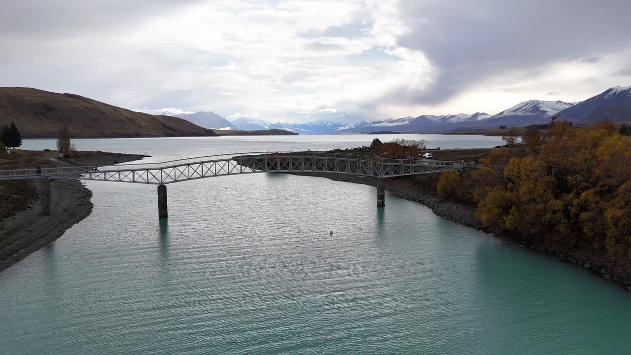 Aerial footage captures a bridge spanning Lake Tekapo with surrounding autumn foliage and mountainous backdrop under cloudy skies
