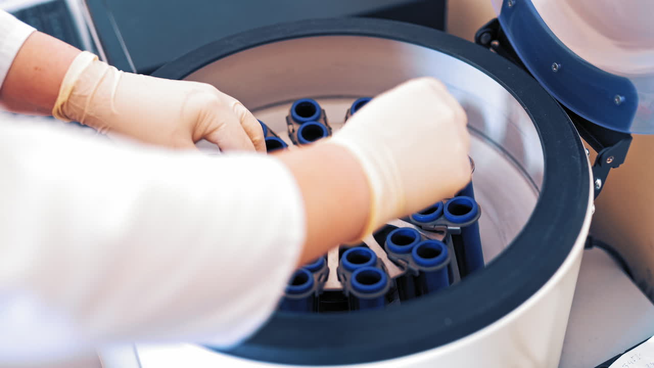 Female scientist loads some vials of liquid into a centrifuge in a laboratory. Close-up hands in protective gloves of a female placing blood samples in the special machine and switches it on.