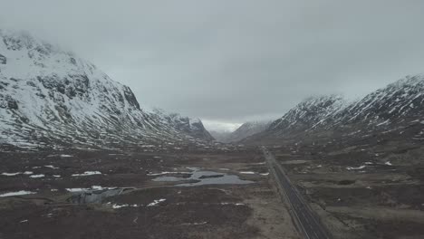 Toma-Aérea-Hacia-Atrás-De-Una-Carretera-De-Montaña-Con-Tráfico-Durante-Un-Día-Nevado-De-Invierno-En-Las-Tierras-Altas-De-Escocia