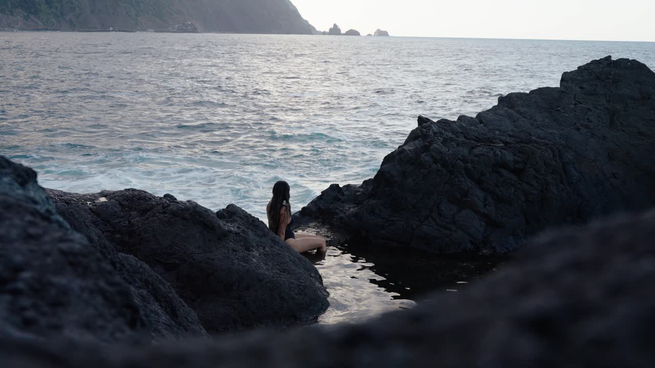 Woman in black swimsuit sits quietly among black rocks, watching the sea and distant cliffs in Seixal, Madeira.