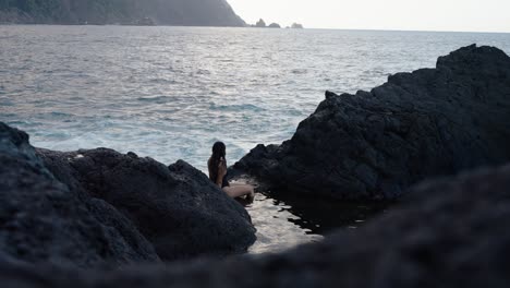 woman in black swimsuit sits quietly among black rocks, watching the sea and distant cliffs in seixal, madeira.