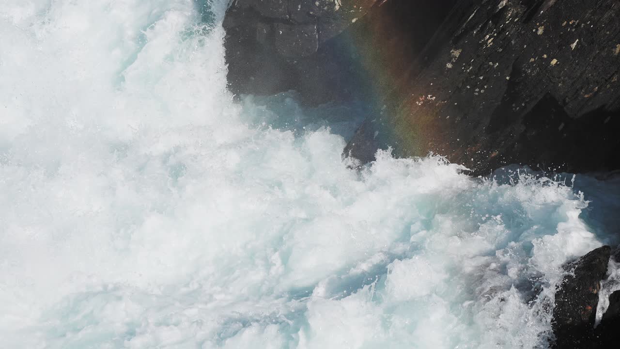 A dynamic scene of turbulent whitewater splashing over the dark rocks, highlighted by a faint rainbow emerging in the water mist