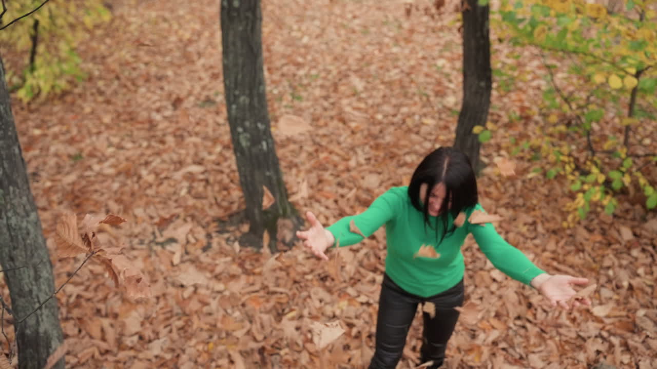 Seorang wanita ber-sweater hijau menyendok dan melemparkan daun-daun kering, close up tangan menyapu tumpukan lalu berdiri dengan tangan terangkat di antara batang pohon oak, celana kulit, sepatu bot di lantai hutan yang renyah, dedaunan cokelat hangat