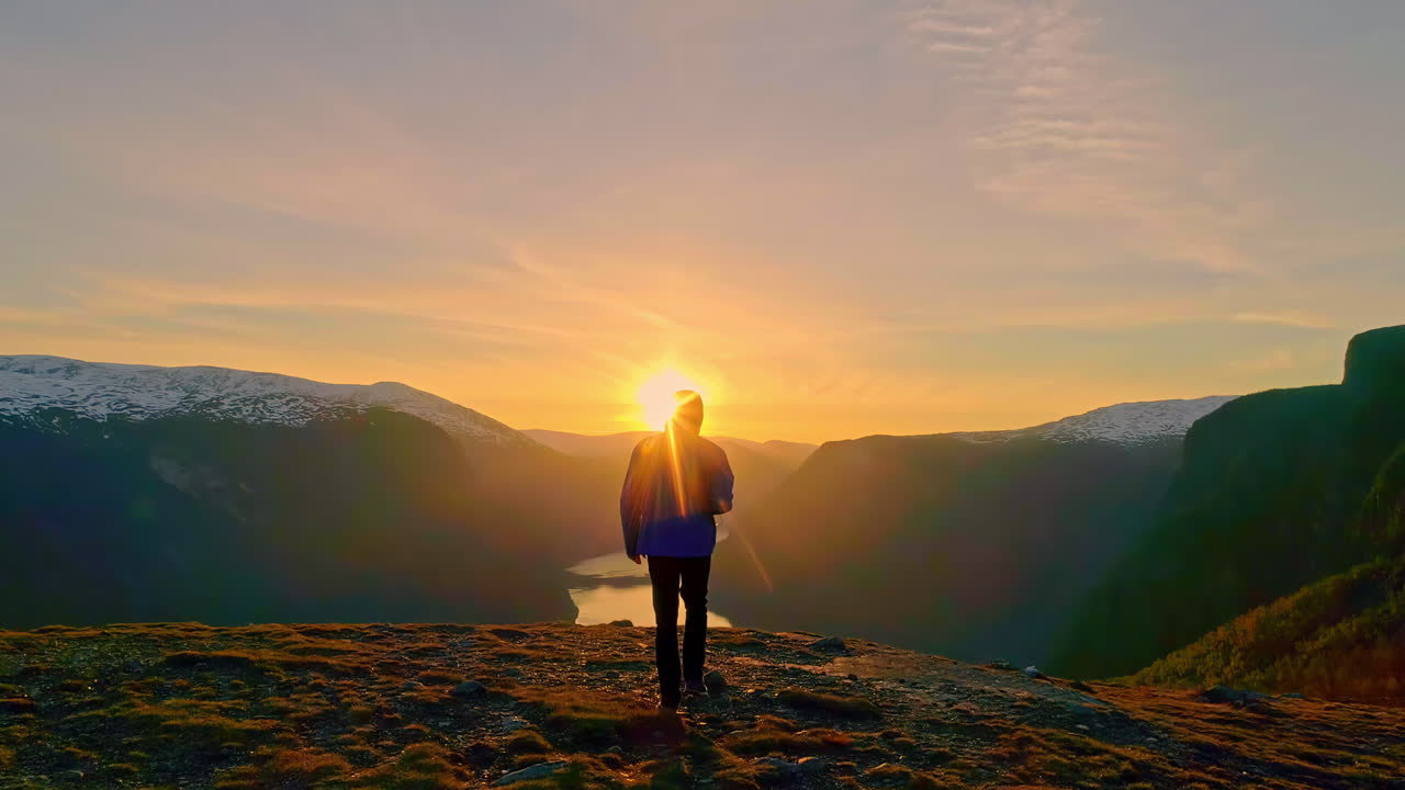 Escursionista con giacca con cappuccio che cammina in cima a una montagna che domina un fiordo all'alba.