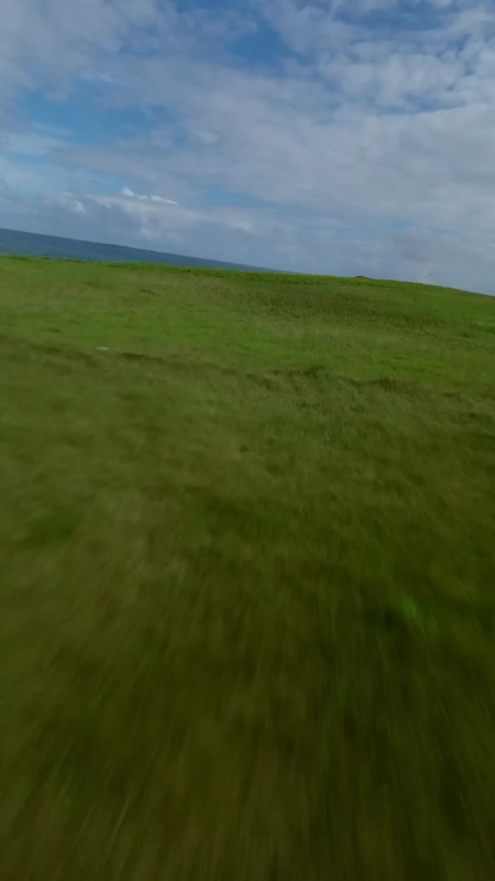 FPV drone rushes toward Atlantic horizon in Brittany, France green cliffs rise above turquoise waves with grassy paths leading to rocky shore under partly cloudy sky