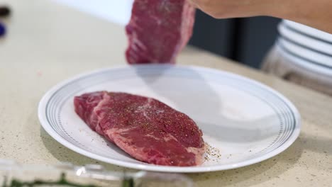 hands seasoning beef steaks with salt and pepper on a plate in a bright kitchen setting