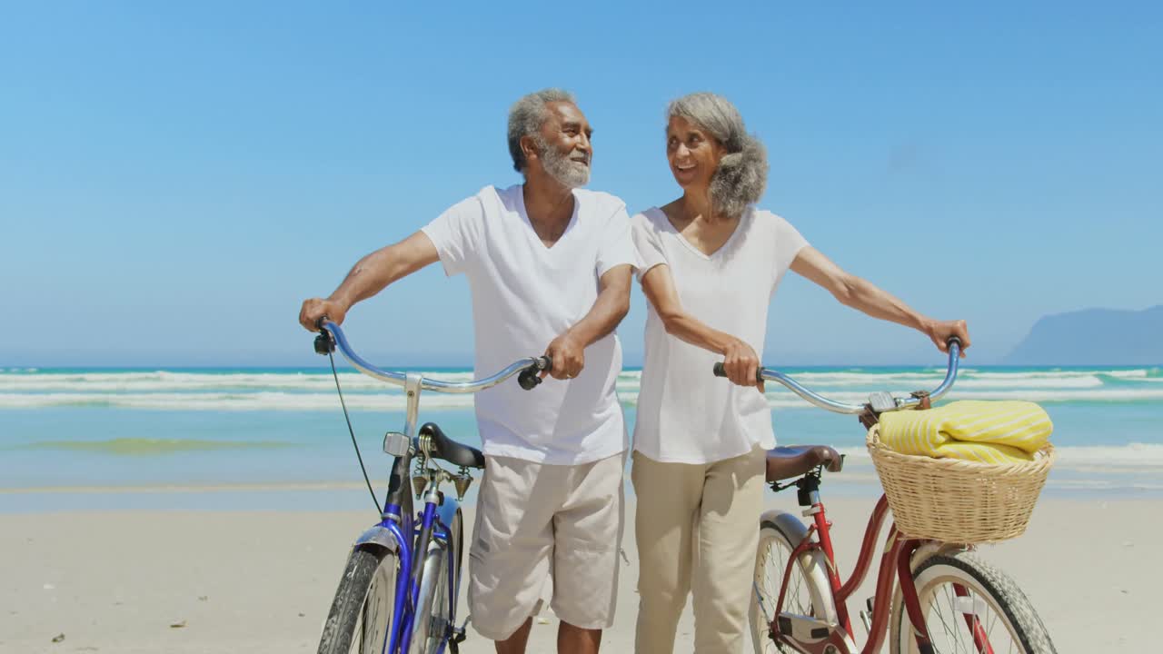 Front view of active senior African American couple with bicycle walking on beach in the sunshine 4k
