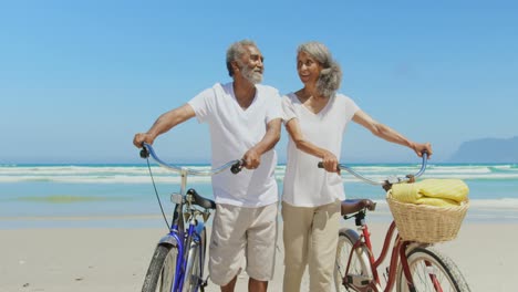 Vista-Frontal-De-Una-Pareja-Afroamericana-Senior-Activa-Con-Bicicleta-Caminando-Por-La-Playa-Bajo-El-Sol-4k