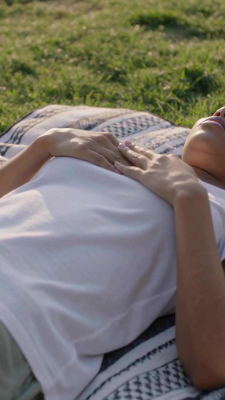 Vertical video: Pulling camera revealing woman lying on blanket keeping hands on chest in white tee