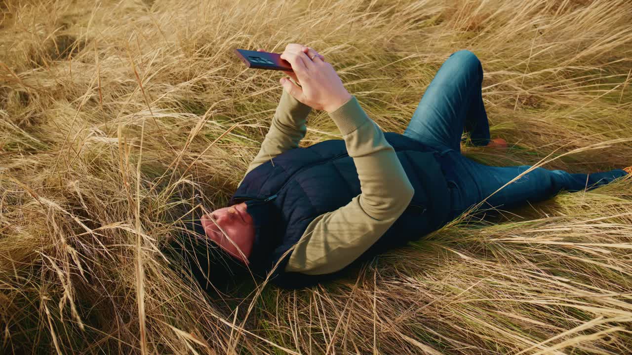 Eine Person entspannt auf einem grasbewachsenen Feld, liegt da und benutzt ein Smartphone, umgeben von hohem, goldenem Gras in einer ruhigen Umgebung im Freien. Fängt die Essenz von Freizeit und Technologie in der Natur ein