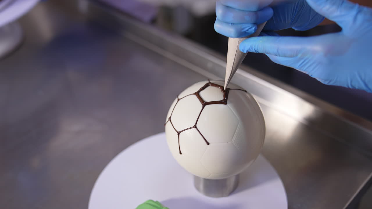 Making cake in a shape of soccer ball. Confectioner's hands applying chocolate lines on the spherical cake. Close up.