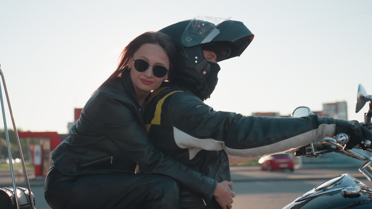 Woman walks up to parked motorcycle, climbs onto back seat, hugs husband rider closely and rests head on his shoulder, both wearing leather jackets, preparing for warm evening road journey together