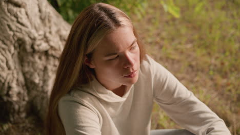 young lady sitting on ground under tree with hand resting over knee, focusing intently on something, dry foliage scattered around adds to natural setting