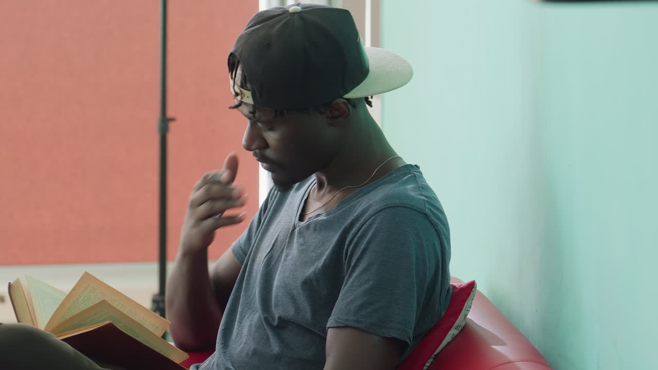 Vista lateral de um jovem estudante do sexo masculino com uma camiseta casual e um boné virado para trás, lendo um livro aberto, tocando a barba pensativamente enquanto segura a página, sentado em um assento vermelho perto de uma parede verde-água