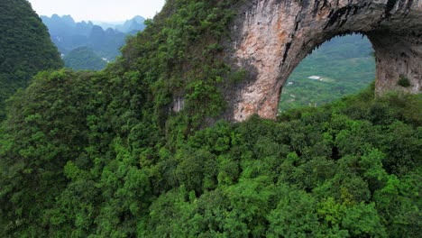 Vista-Aérea-Del-Arco-De-Moon-Hill-En-El-Exuberante-Yangshuo-Con-Una-Persona-Explorando-Debajo,-China