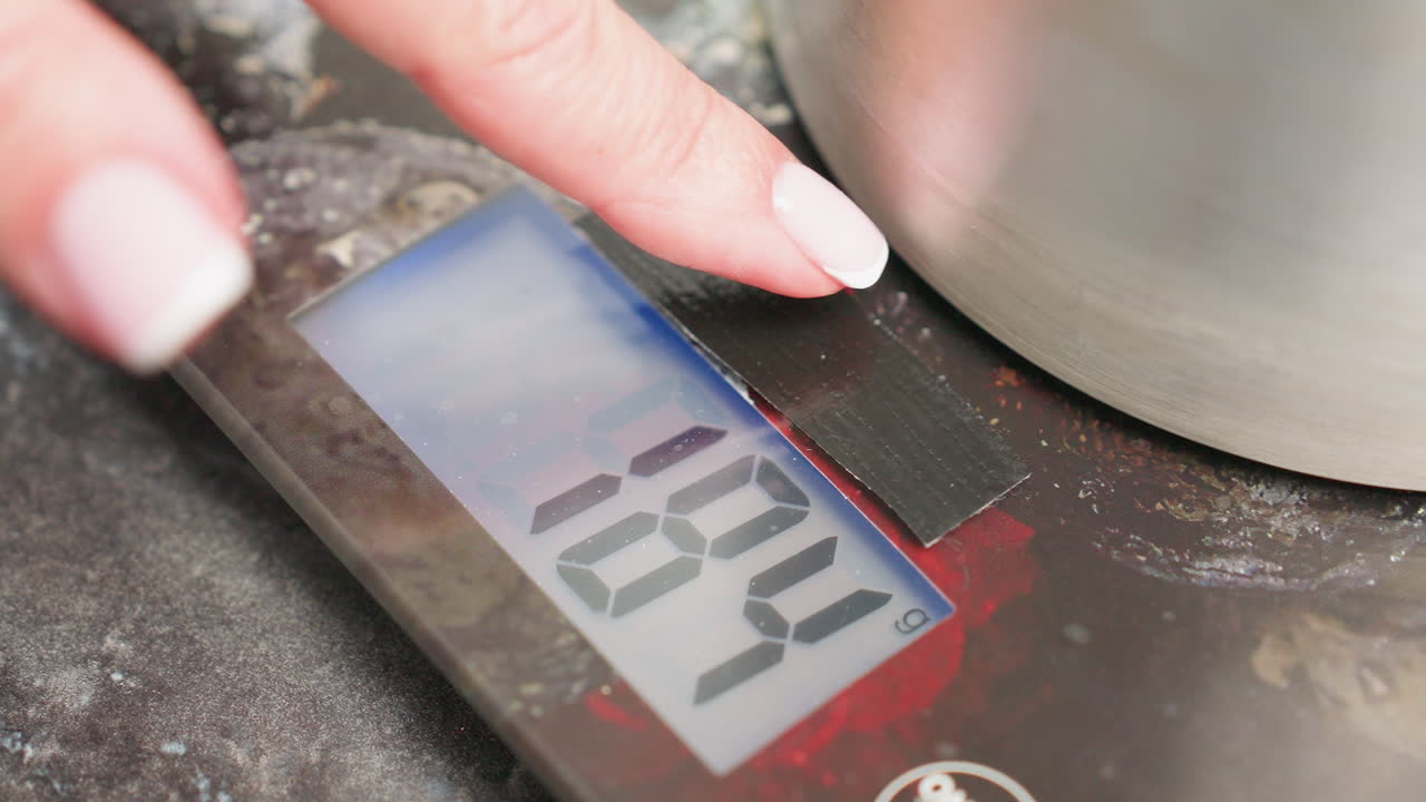 Close up of woman hand with manicured nails pressing tare button on digital kitchen scale while metal container rests on surface, resetting measurement to zero for accurate weighing