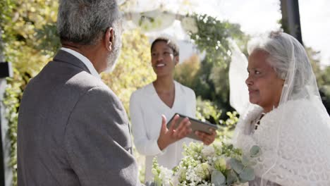 mujer afroamericana oficiando la ceremonia de boda de una pareja biracial mayor en el jardín, cámara lenta