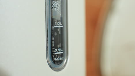 Close up of electric kettle water level indicator showing bubbles and steam rising inside during boiling process, highlighting transparent gauge markings in bright kitchen