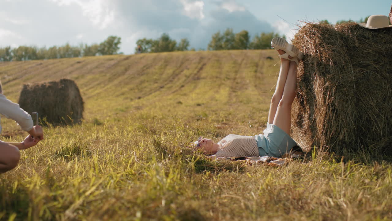 dama en cuclillas tomando fotos de su hermana al aire libre mientras coloca las piernas en el heno en vastas tierras de cultivo, cálido entorno rural con campos dorados, atmósfera rústica y luz solar que arroja sombras suaves