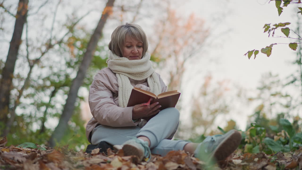 middle age woman wearing warm scarf and jacket seated cross-legged on leafy ground flipping through book pages calmly, immersed in peaceful nature with soft morning light filtering through trees