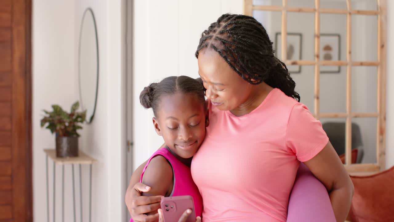 Mãe africana com top coral e filha reagindo a alerta de telefone, rindo e se abraçando em casa com tapete.