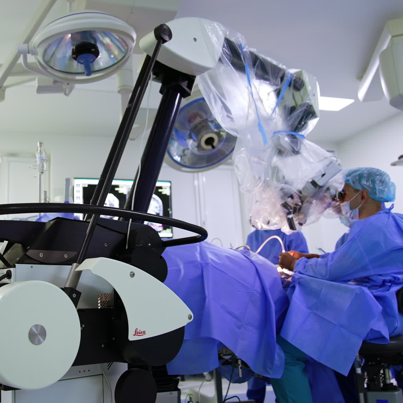 Advanced technological microscope standing on the floor of modern surgery room. Doctor sits at the patient performing operation and looking at microscope.