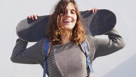 Portrait-of-happy-mixed-race-woman-holding-skateboard-behind-head-smiling-in-the-sun