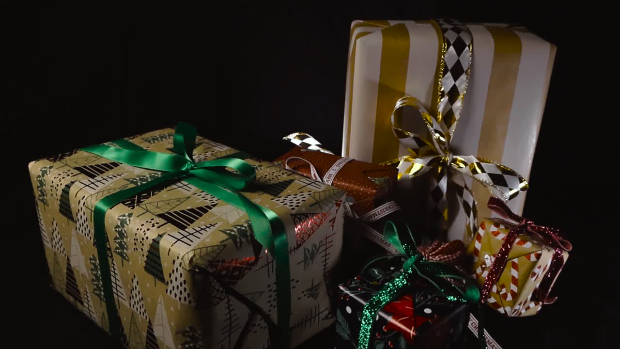 Primer plano con movimiento suave sobre regalos de Navidad envueltos y colocados sobre un fondo oscuro en un estudio con lazos festivos alrededor de las cajas de regalo y un elegante resaltado para la promoción de la temática navideña y espacio para texto.
