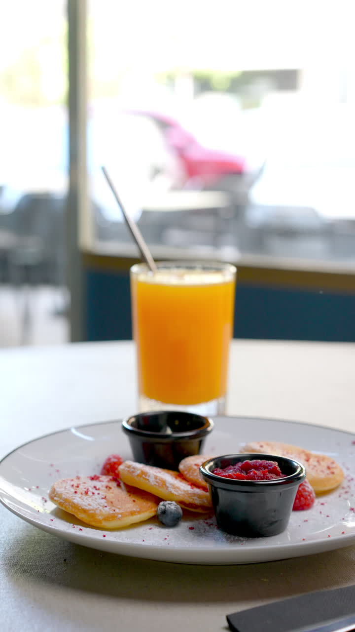 Small pancakes with powdered sugar served with fresh berries and jam, accompanied by a glass of orange juice, compose a delightful breakfast on a white table in a cafe