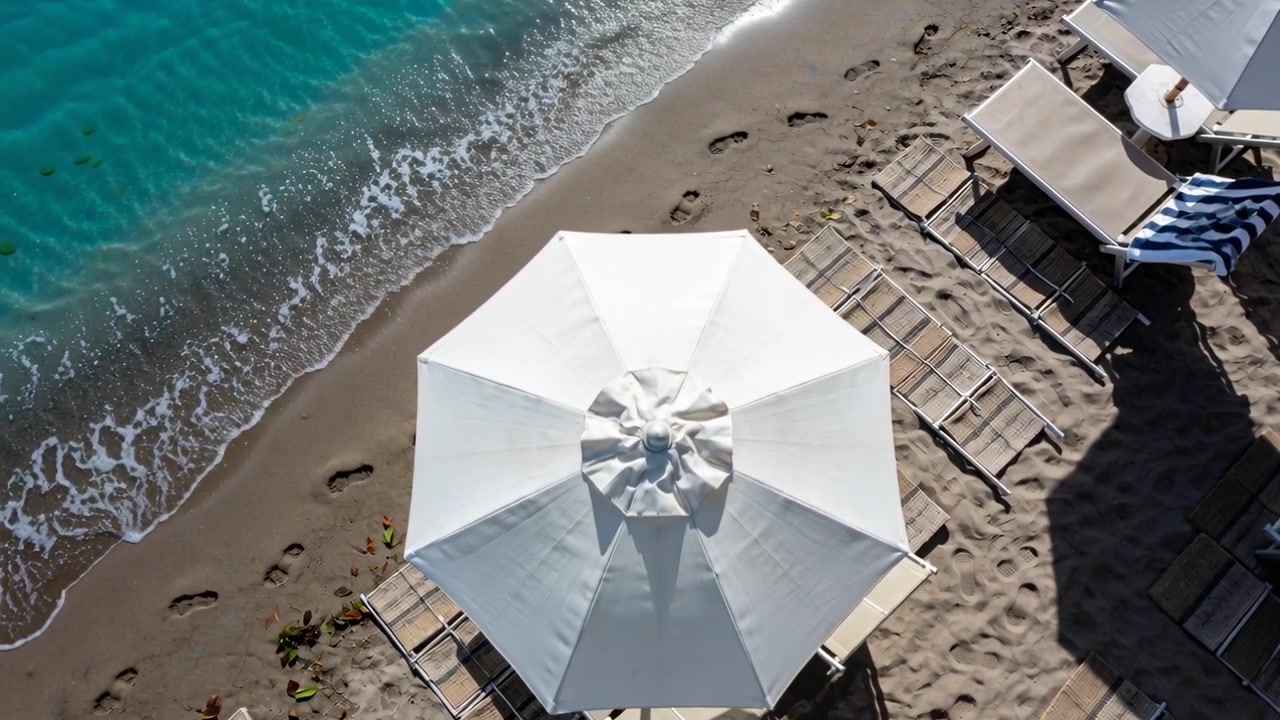 Drone in volo che rivela la costa sulla spiaggia, mostrando un ombrellone bianco, lettini e impronte.