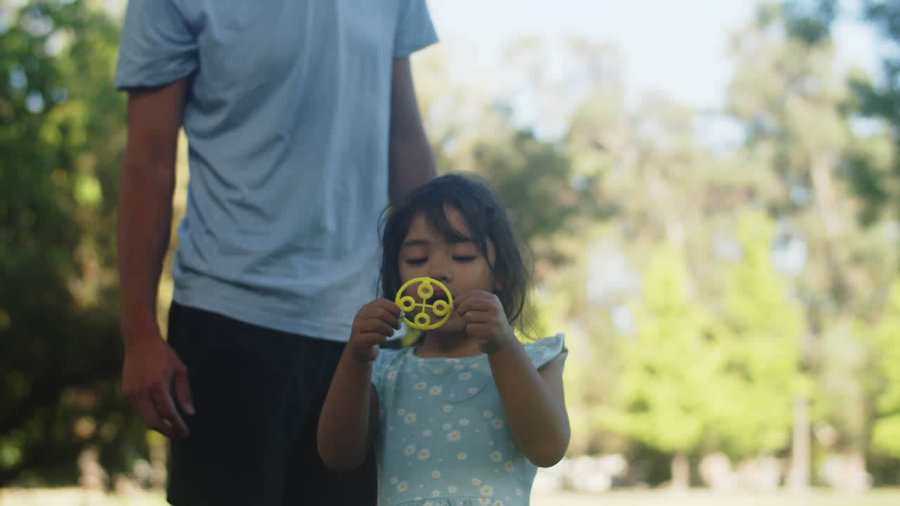 Cute Asian little girl blowing soap bubbles with father in park