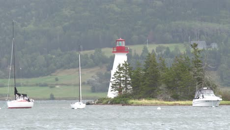 it is a beautiful lighthouse with 3 sailboats in the water with a lot of trees behind it