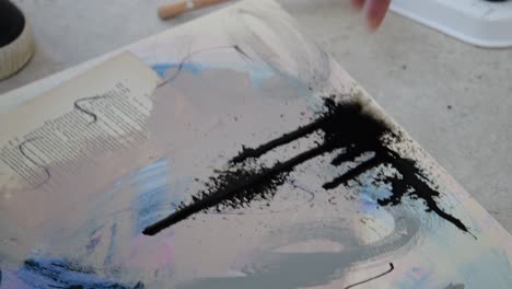 Female-artist-in-studio-holds-canvas-with-ink-splatter,-spraying-it-with-water-to-create-dripping-effect