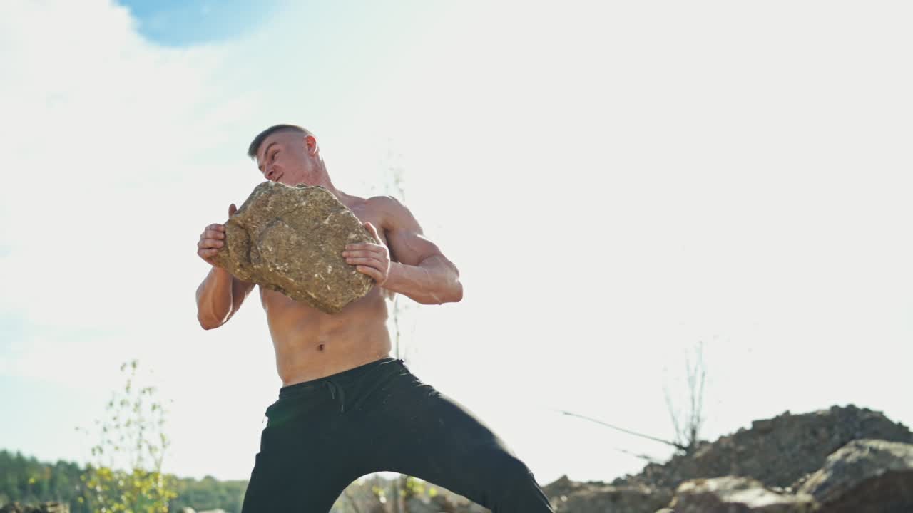Muscular man with trained body exercises with stones. Strong bodybuilder doing workout by throwing heavy rocks on nature background.