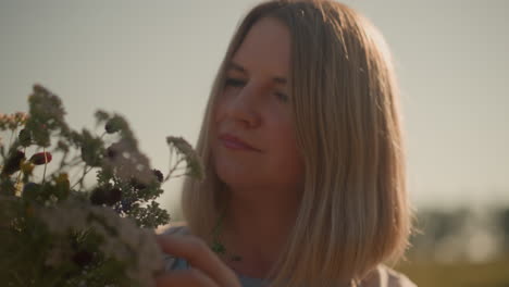 close-up of woman smiling while enjoying scent of freshly picked wildflowers in vast farmland, holding vibrant floral bouquet with soft blurred distant trees in background